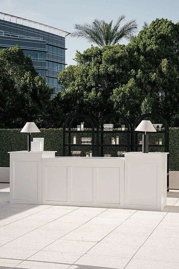 Wedding bar setup with a white bar counter, arched shelves of glassware, and votive candles against a hedge wall on an outdoor patio