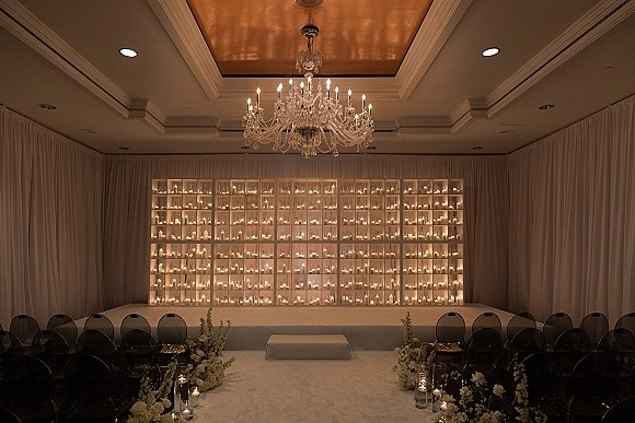 Ceremony setup with wedding candle wall backdrop, white aisle runner lined with glass votive candles and white roses in an indoor ballroom