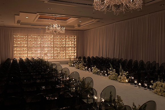 Indoor ceremony setup with a ballroom wedding ceremony candle wall backdrop, white draping, chandelier, and aisle lined with candles and flowers