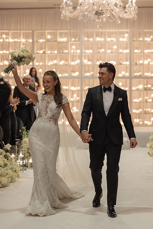 Wedding recessional as bride and groom walk the aisle holding hands, bride raising bouquet beneath a chandelier, candle wall and guests applauding