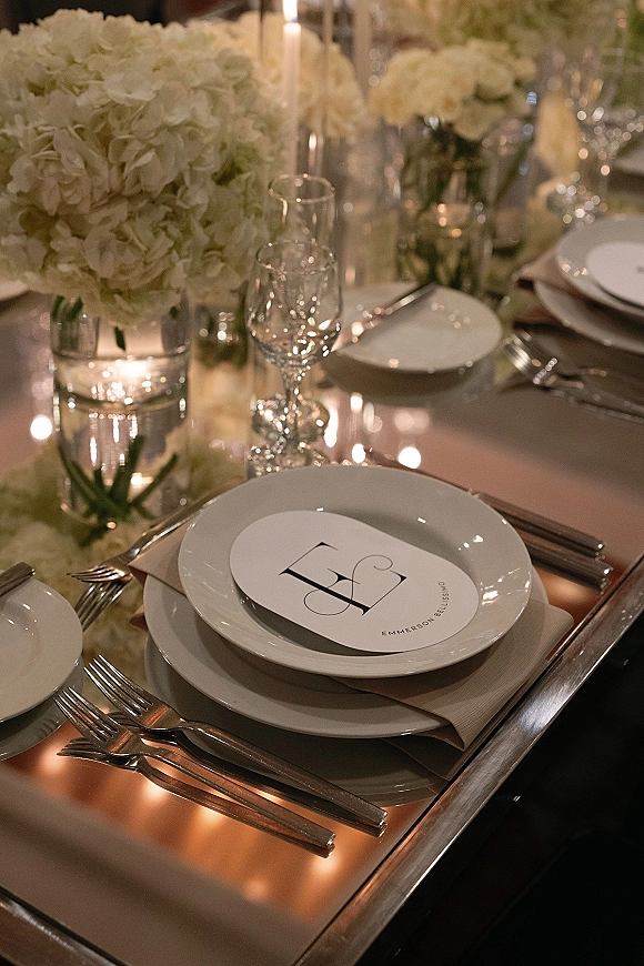 Reception tablescape with wedding place setting, white hydrangea centerpiece and taper candles on a copper tabletop in warm candlelight