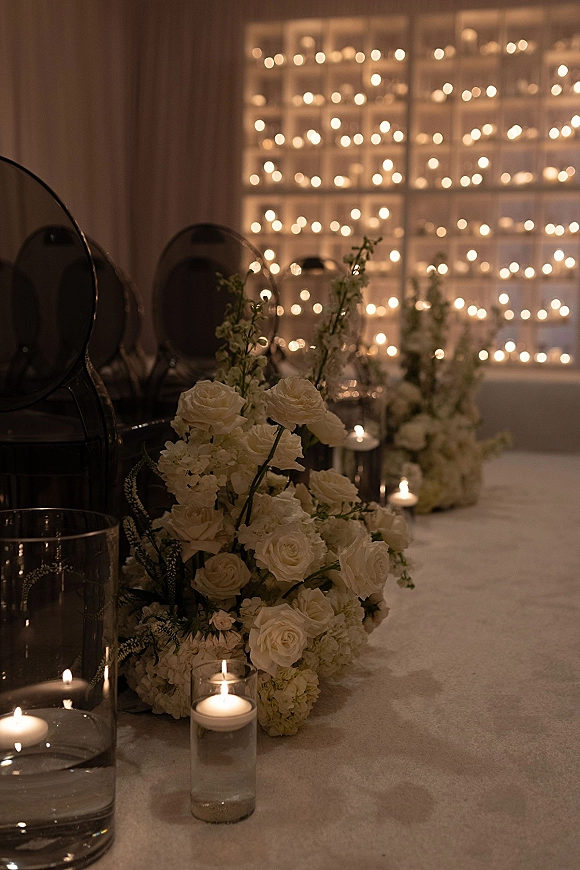 Reception tablescape with a wedding candle centerpiece of white roses and hydrangeas, greenery, and glass votives under warm string lights and drapery