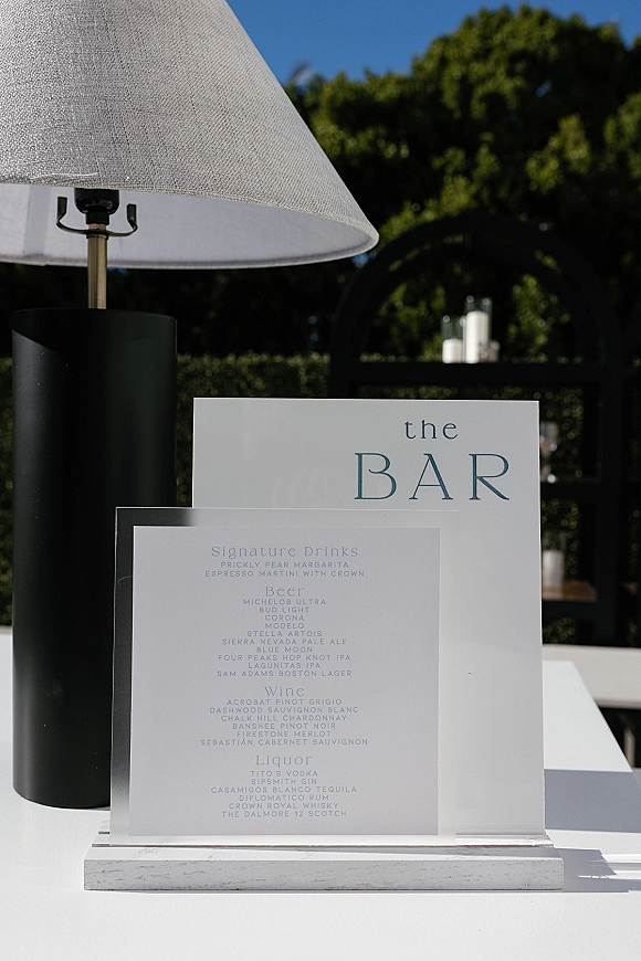 Wedding bar sign on clear acrylic with serif drink menu beside a table lamp on a white tabletop, set against outdoor greenery and blue sky