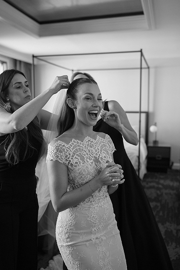 Bridal getting ready as a bridesmaid helps the bride putting on veil over an off-the-shoulder lace gown in a window-lit bedroom