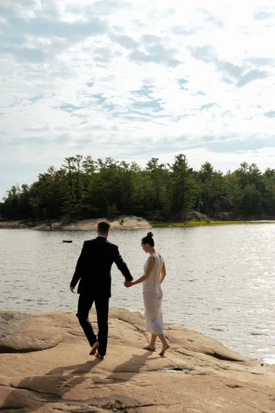 Couple portrait of bride and groom walking along a rocky lakeshore, holding hands, her bridal gown moving softly by pine trees under a cloudy sky.