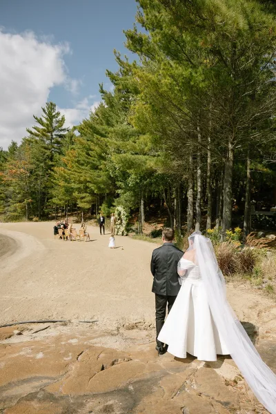 Wedding processional with a bride walking down the aisle in a long veil amid pine trees and a sandy aisle under blue sky.