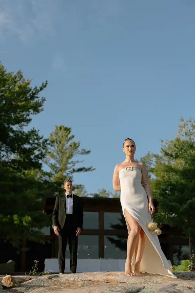 Couple portrait of a groom and a bride with roses, a simple headband catching light near pine trees and a cabin exterior with glass windows.