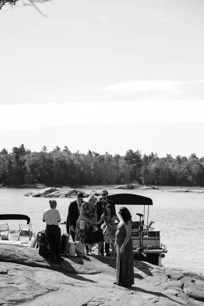 Wedding guests arrive by boat at a lakeside venue, stepping onto a rocky shoreline as trees frame the water beneath a wide open sky.