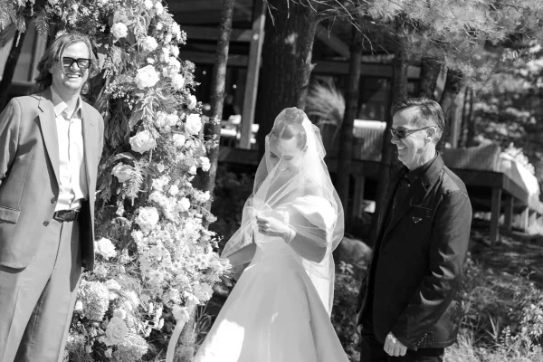 Ceremony moment with a bride adjusting her veil beneath a floral arch in a garden framed by trees and a rustic wooden structure.