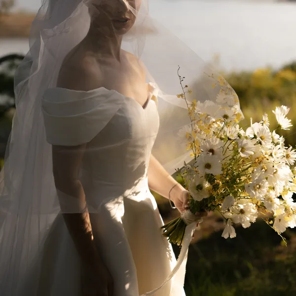 Bridal portrait of a bride with veil, standing by a calm lakeside shoreline amid grass and foliage in natural light.