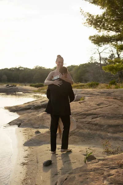 Couple portrait of a bride and groom embrace on a rocky shoreline beach, her wedding ring catching light as waves lap and trees frame the water.