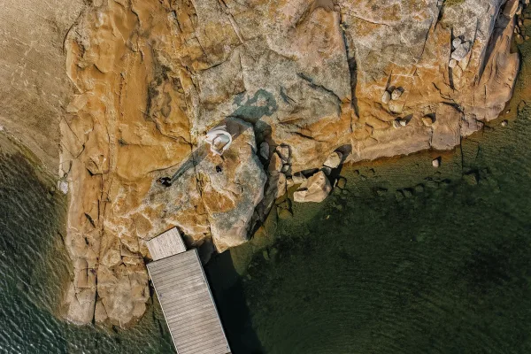 Bridal portrait of a bride on rocks, holding a bouquet beside a wooden dock on a rocky shoreline with boulders along the water.