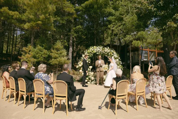 Ceremony moment as the couple exchange vows in an outdoor wedding ceremony beneath pine trees, framed by a floral arch on sandy forest ground.