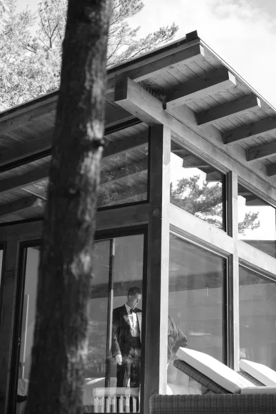Black and white groom portrait of a groom in tuxedo with a neat bow tie, framed by glass windows and wood beams with trees visible beyond.