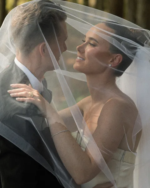 Couple portrait with a veil over the couple as they embrace, the bride gazing at the groom amid forest trees in soft natural light.