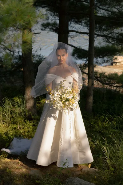 Bridal portrait of a bride with veil standing beside a tranquil lake, trees and grass softly blurred in the background
