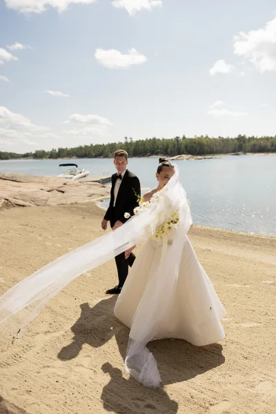 Couple portrait, a lakeside wedding portrait with the bride's flowing veil as they walk the sandy beach past a dock and calm lake.