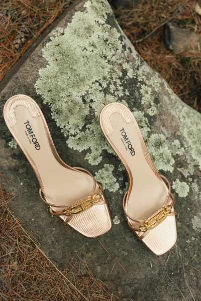 Bridal shoes in rose gold with gold chain hardware rest on a lichen-dusted rock scattered with pine needles.
