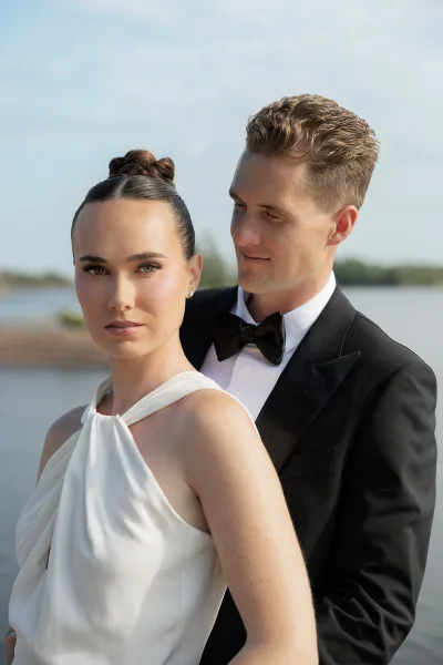 Couple portrait as a bride and groom embrace by the water, the groom's bow tie crisp, with open sky, a calm shoreline, and distant trees behind.