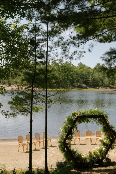 Ceremony setup centered on a hexagon arch, styled for lakeside ceremony decor at a sandy shore framed by pine trees beneath an open sky.