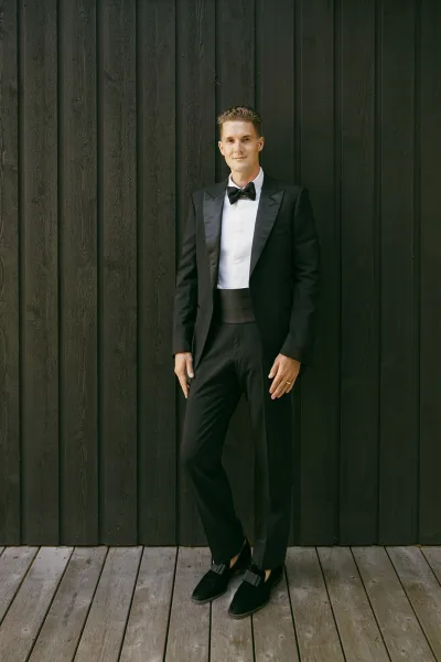 Groom portrait of a black tie groom with satin lapels, standing against a dark wood wall on a wooden deck, hands relaxed and expression confident.