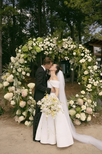 Wedding kiss portrait capturing an outdoor couple kissing beneath a circular floral arch, framed by trees on sandy ground in a quiet forest.