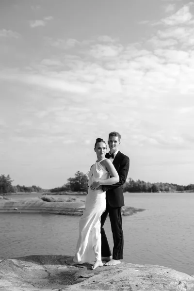 Couple portrait of a barefoot bride and groom on a rocky shore, a lakeside wedding portrait with rippling water, distant trees, and a cloudy sky.