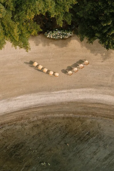 Beach ceremony setup with lakeside ceremony seating on a sandy shoreline, framed by low ground florals beside calm water and trees beyond.