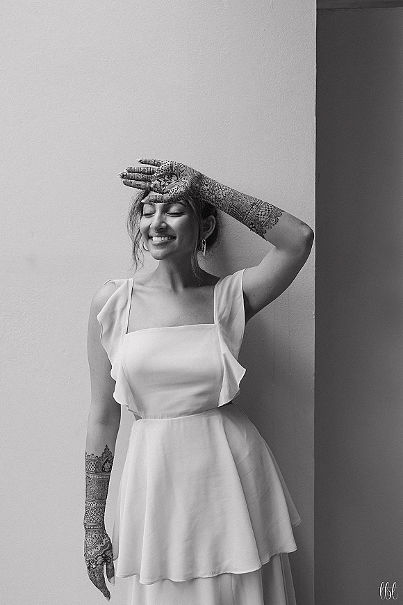 Bridal portrait of a bride showcasing bridal mehndi on her arm, wearing a short white square-neck dress and hoop earrings by a plain wall