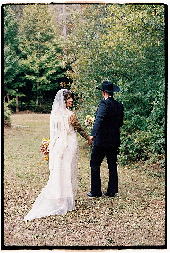 Couple portrait of bride and groom holding hands from behind, groom in cowboy hat and suit, bride in veil with bouquet in woods