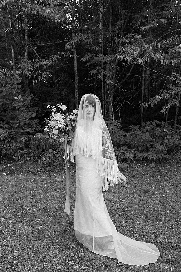 Bridal portrait in black and white of a bride holding bouquet, wearing a fringe veil and satin dress, standing among forest trees