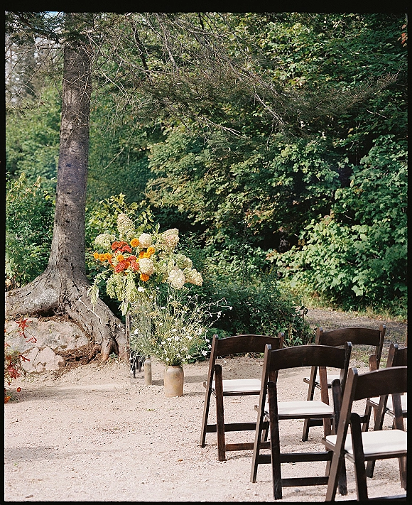 Ceremony seating with outdoor wedding chairs, wood folding chairs and cushions lining a forest aisle with orange and white flowers