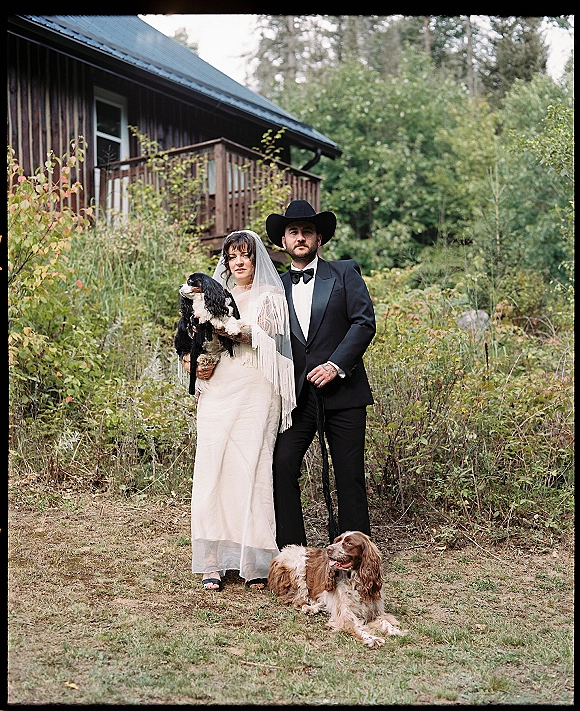 Couple portrait of bride in veil and fringe shawl with groom in tuxedo and cowboy hat holding wedding dogs on a cabin porch