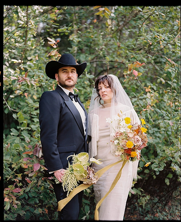 Couple portrait in a forest wedding portrait, bride in veil and fringe shawl beside groom in cowboy hat holding bouquets amid trees