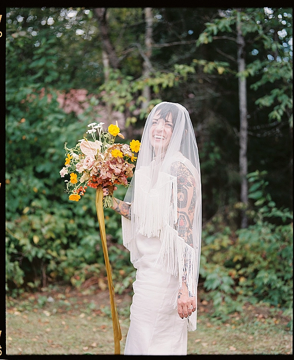 Bridal portrait of a bride holding bouquet, laughing with a fringe veil over her face, in a green garden with trees behind