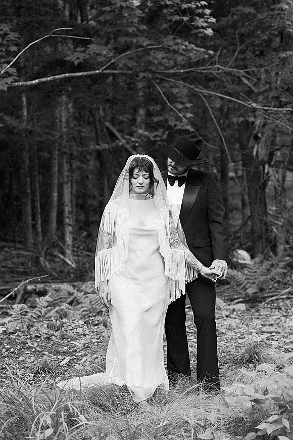 Couple portrait in a black and white wedding portrait style, holding hands in a forest, bride in fringed veil and groom in cowboy hat