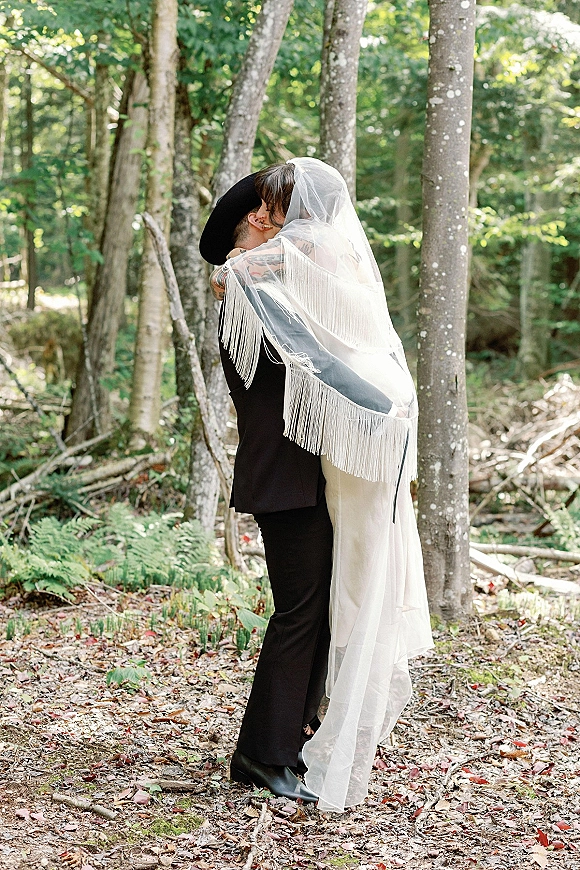 Wedding hug as the groom in a wide brim hat lifts the bride in a fringe shawl, their veil draped over them amid forest ferns