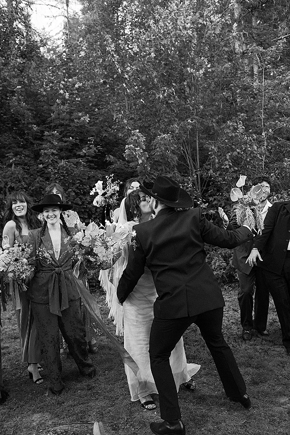 Wedding kiss in a black and white wedding photo as the bride in veil and groom in wide brim hat walk with cheering party in a garden lawn