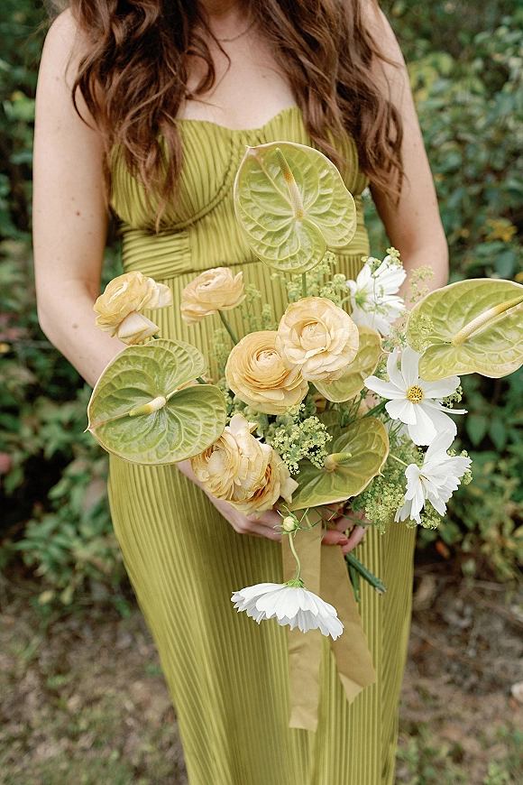 Bridesmaid bouquet with anthurium wedding bouquet leaves, yellow ranunculus and white cosmos, held against a green pleated dress outdoors