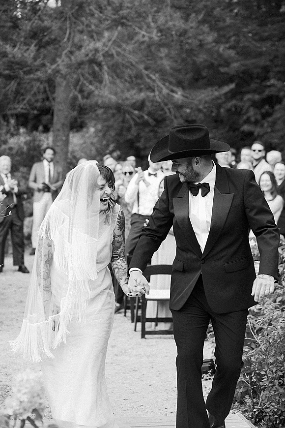 Wedding recessional as newlyweds walk down the aisle holding hands, bride in fringe veil and groom in cowboy hat amid cheering guests outdoors