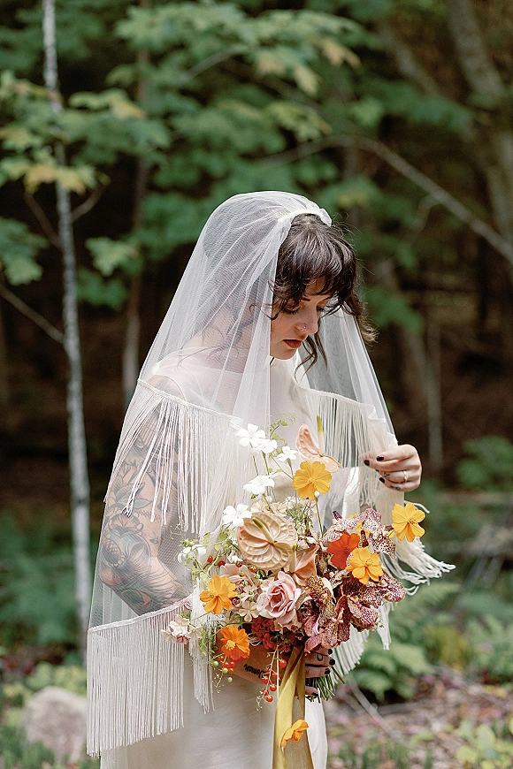 Bridal portrait of a tattooed bride holding a statement bouquet with orchids and roses, her fringe veil flowing in a forest backdrop