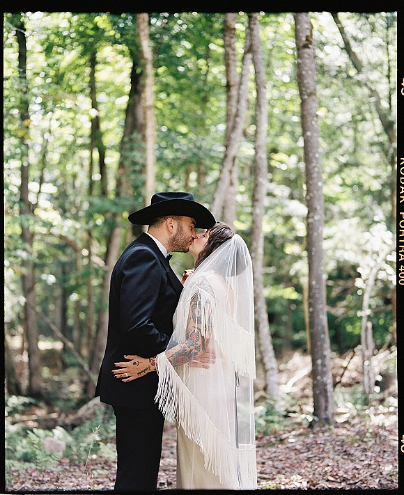 Wedding kiss portrait of bride and groom kissing in a forest, her fringe veil flowing as he wears a cowboy hat with tuxedo