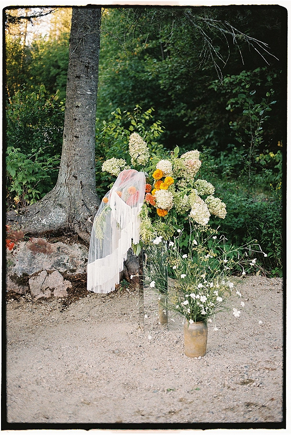 Wedding florals and ceremony floral installation with hydrangeas and orange blooms in glass vases, draped veil, set on a forest floor
