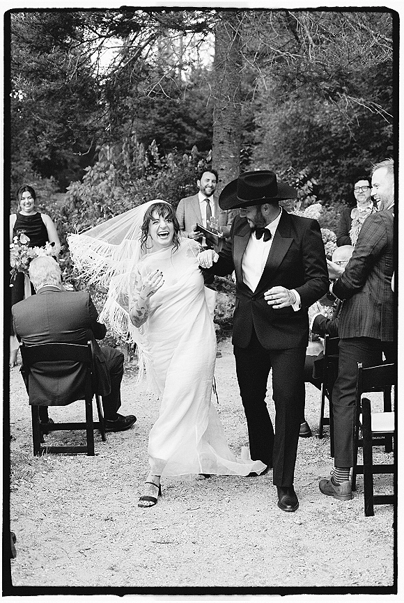 Recessional moment as newlyweds walk the aisle, bride in fringe veil laughing beside groom in cowboy hat, guests cheering on garden walkway
