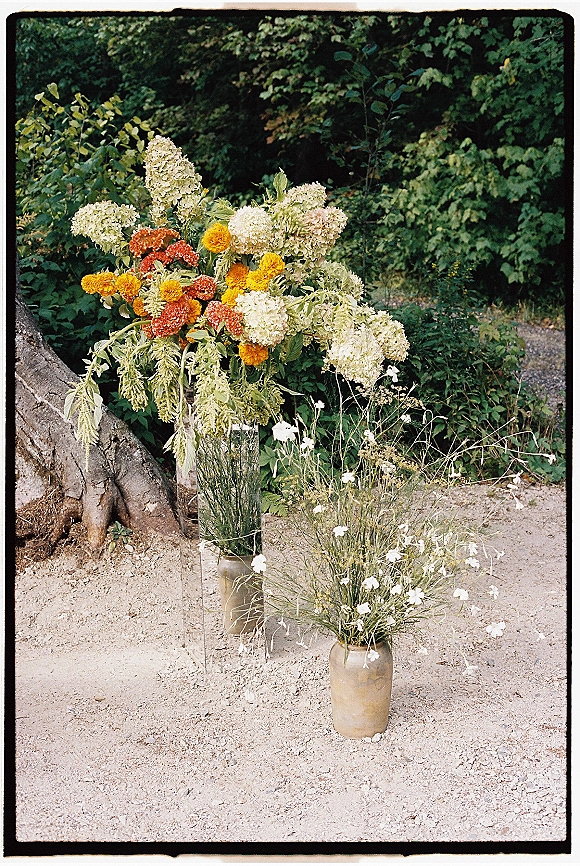 Wedding floral arrangements in ceramic vases with wildflower stems, white hydrangea and marigold blooms on gravel beside garden shrubs and trees