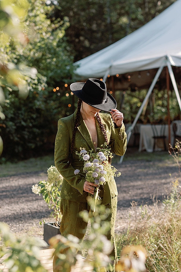 Bridal portrait of a bride in suit wearing a green bridal suit and wide brim hat, holding a bouquet on a gravel path by a tent with string lights