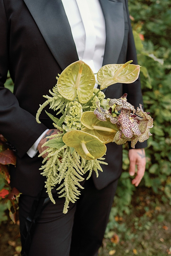 Bridal bouquet with anthurium and orchids cascading with greenery, held against a groom’s black tuxedo on a garden walkway outdoors