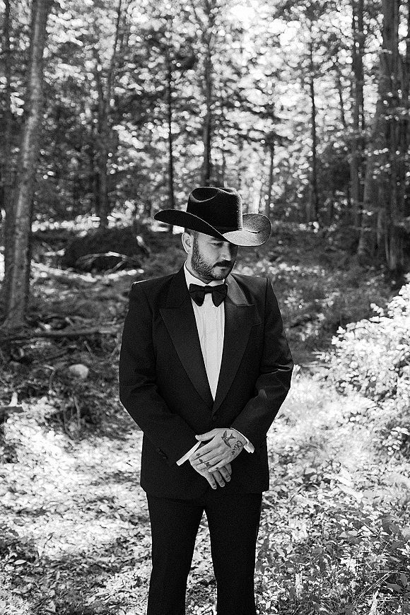 Groom portrait wearing a cowboy hat and black tuxedo with bow tie, standing hands clasped on a wooded path in dappled sunlight