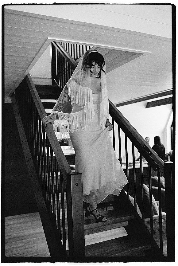 Bridal portrait of a bride on staircase in a satin dress with fringe veil and heels, showing tattoos, indoors with guests seated behind
