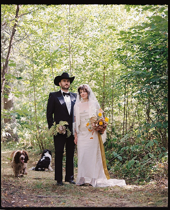 Couple portrait in a forest, bride in wedding dress and veil with bouquet beside groom in tuxedo and cowboy hat, dogs on path